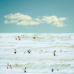 Billowy white clouds float in a turquoise blue sky above a white ocean as swimmers and surfers play in the cresting waves.