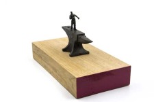 A heroic figure in bronze holds a hammer to his chest, standing on an oversized anvil moutned on a block of wood painted pink on the edges. Image 3