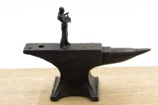 A heroic figure in bronze holds a hammer to his chest, standing on an oversized anvil moutned on a block of wood painted pink on the edges. Image 6